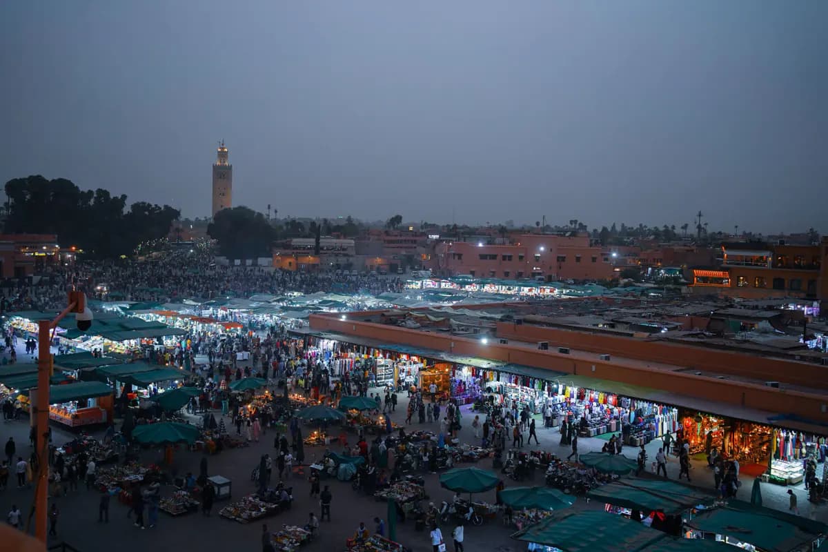 Jemaa el-Fna at dusk seen from above, crowded with lit stalls, with the Koutoubia minaret in the distance