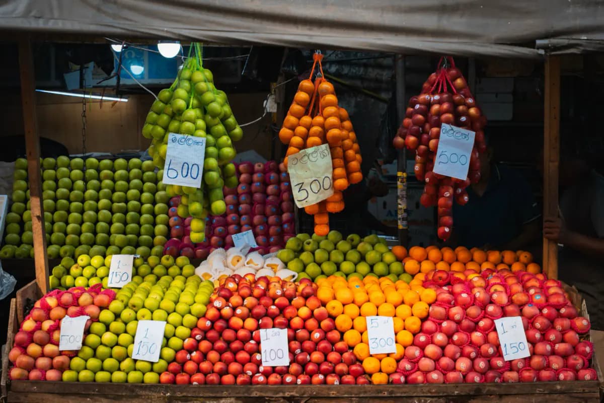 Apples and oranges stacked high on a market stall in Colombo, with more fruit hanging in nets and handwritten cards