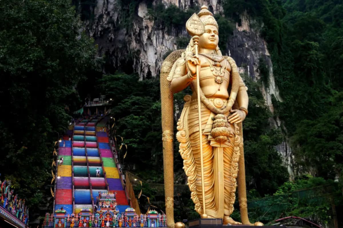 The golden statue of Murugan beside the painted steps at Batu Caves, below a limestone cliff covered in trees