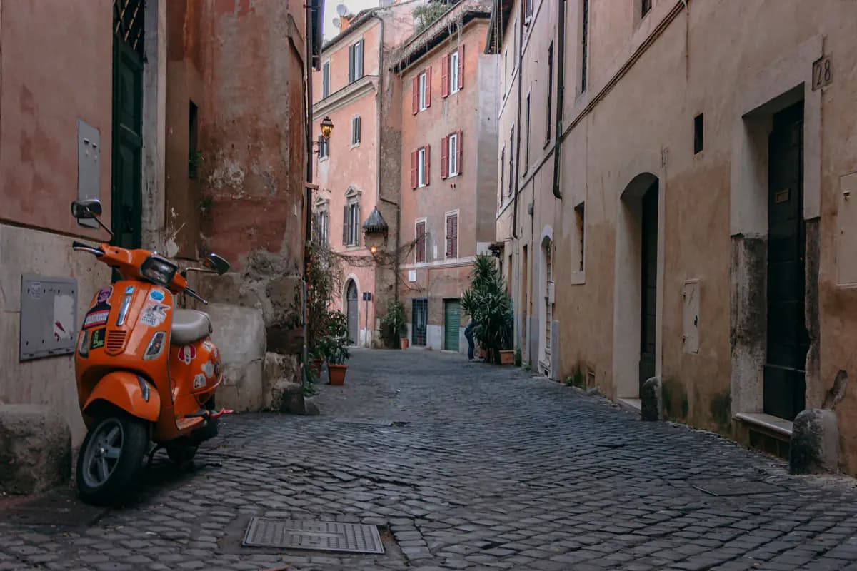 An orange scooter parked on a narrow cobbled street in Trastevere, between old ochre and pink houses with shutters