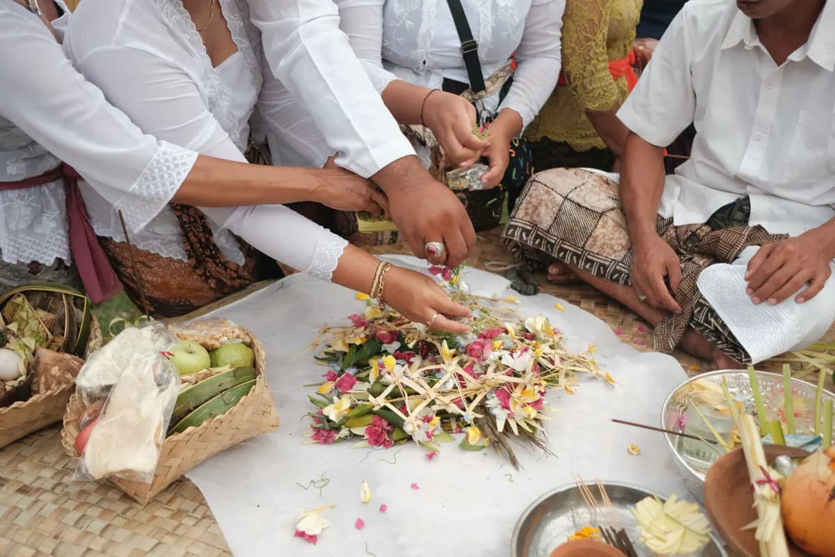 Hands arranging flowers and woven palm leaf offerings on a white cloth beside baskets of fruit at a Balinese ceremony