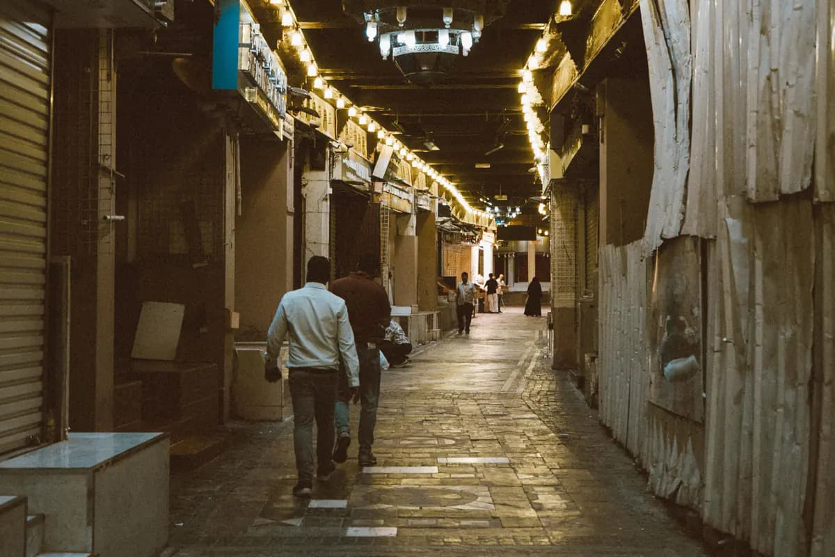 A covered lane of Muttrah Souq lit by strings of lights in the evening, with people walking between shuttered shops